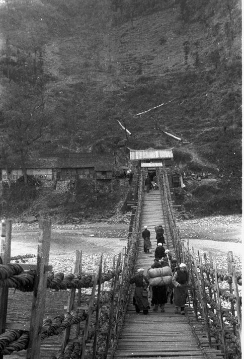 Black and white view of people carrying packs of goods across a bamboo cable suspension bridge over a river near Guanchengzhen