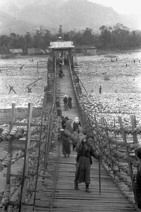 Black and white view of people carrying packs of goods across a bamboo cable suspension bridge over a river near Guanchengzhen