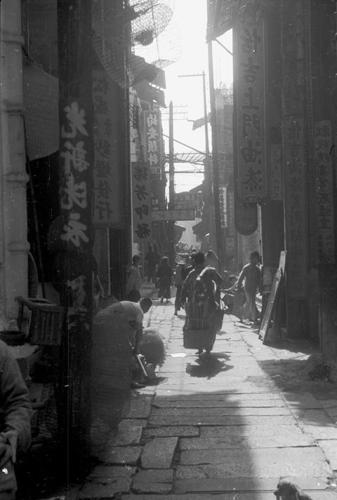 Black and white view of a cobblestoned alleyway in Canton with several people carrying goods and walking into storefronts. Both sides of the alley feature vertical signs with names of shops in Chinese characters. In the background of the view there are electrical poles high above the street.