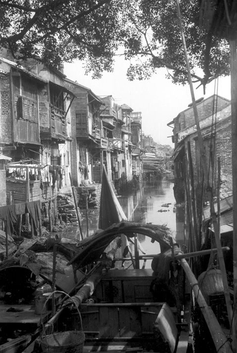 Black and white view from a canal boat of an empty canal in Canton with two-level wooden apartments crowding both sides of the water. There is laundry hanging on lines from the apartments.
