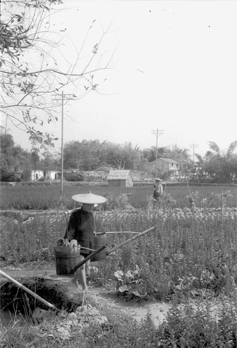 Black and white view of a person wearing a large straw hat carrying two person-sized watering cans in a field. In the background of the view there are electrical poles and several small farm structures and a man with a scythe.