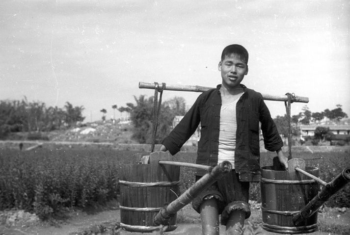 Black and white close-up view of a person carrying two person-sized watering cans in a field.
