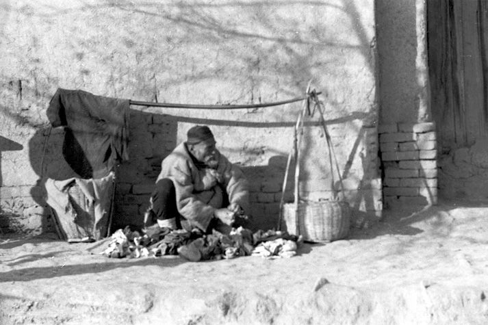 Black and white view of an old person sitting on a dirt road next to a stone gate of Xi'an. They sit between two woven baskets. Above them there is a wooden pole, with their clothing hanging on it.