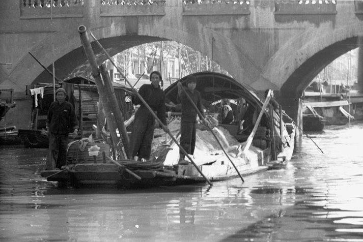 Black and wite view of three people on a boat in the Pearl River transporting goods underneath the West Bridge which leads to the British and French concession of Shamian island. All three people hold long poles used as paddles.