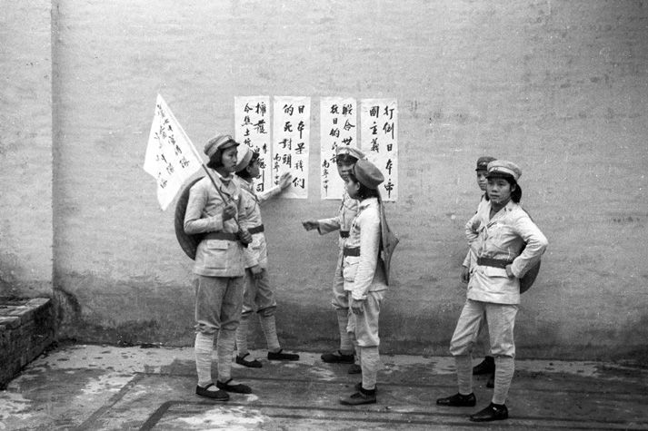 Black and white view of six people wearing light-coloured uniforms, reading four posters on a stone wall outdoors. One person is holding a flag with Chinese characters.