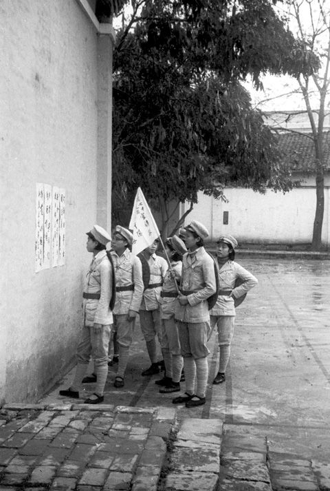 Black and white view of six people wearing light-coloured uniforms, reading a poster on a stone wall outdoors. One person is holding a flag with Chinese characters.