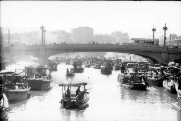 Black and white view of the Zhapu Road Bridge from the water of Suzhou Creek. There are many passenger and merchant boats on the water on either side of the Creek. There is a bus and people walking across the bridge, which has tall street lamps. Behind the bridge there are large buildings in the distance.