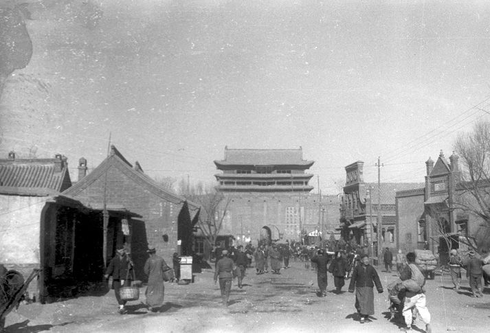 Black and white view of the south gate of the city of Taiyuan. People are carrying goods down a dirt road with stone buildings and electrical poles on either side, leading up to the stone city wall and south gate built in the style of Ming Dynasty architecture.