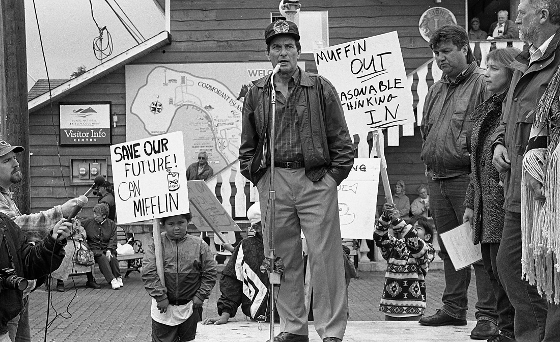 Protestor standing in front of a microphone. Other speakers line up to the protestor's right. To the left, two journalists hold up other microphones. Other protestors can be seen holding signs in the background