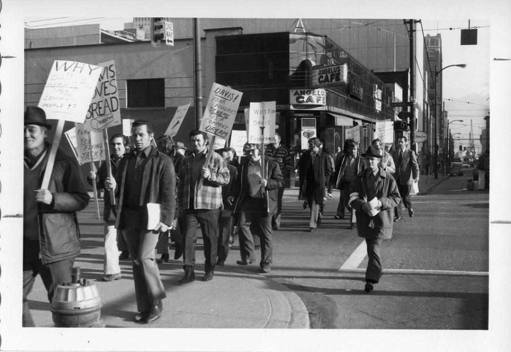 Group of protestors walking down a street in demonstration holding many signs; one sign is in Russian 