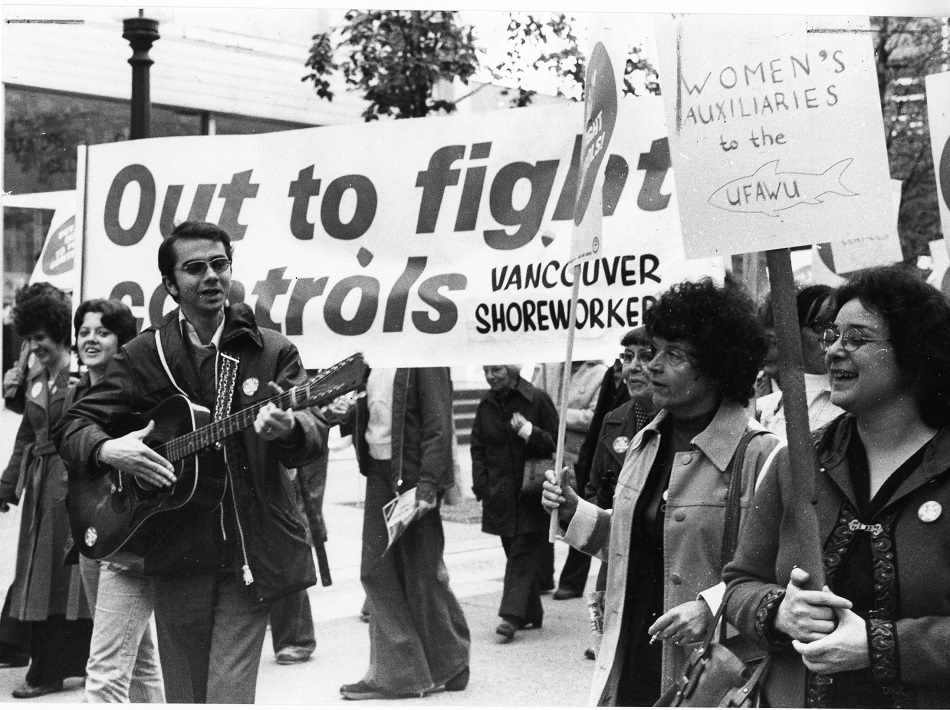 Group of protestors walking with signs. One protestor has a sign in support of the "Women's Auxiliaries to the UFAWU". Another protestor walks with a guitar