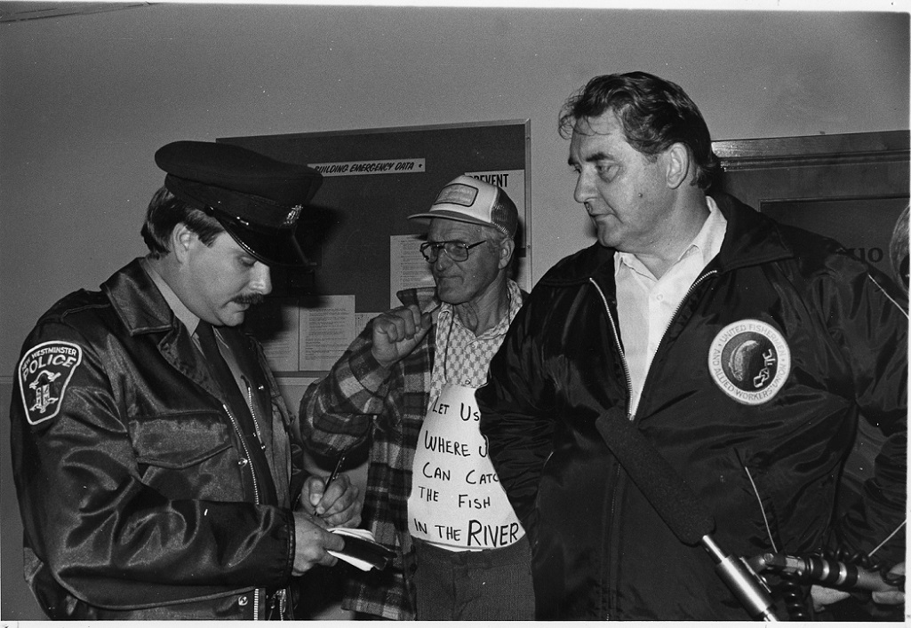 Two protesting fishers stand next to a cop who is writing on a notepad