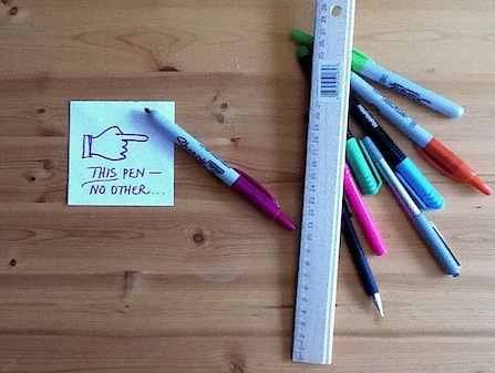 Image shows several pens and a sticky note pointing to one pen in particular. Text on sticky note reads: "THIS pen, no other..." Image demonstrates the grammar rule for non-descriptive adjectives