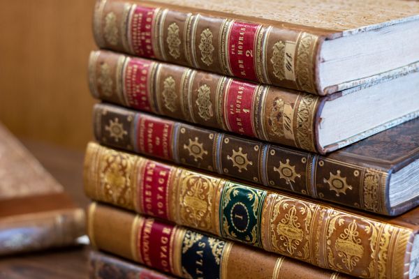 Ornately bound editions from the Anfield Collection stacked on top of each other, including voyages by Cook, Forster, and Mofras