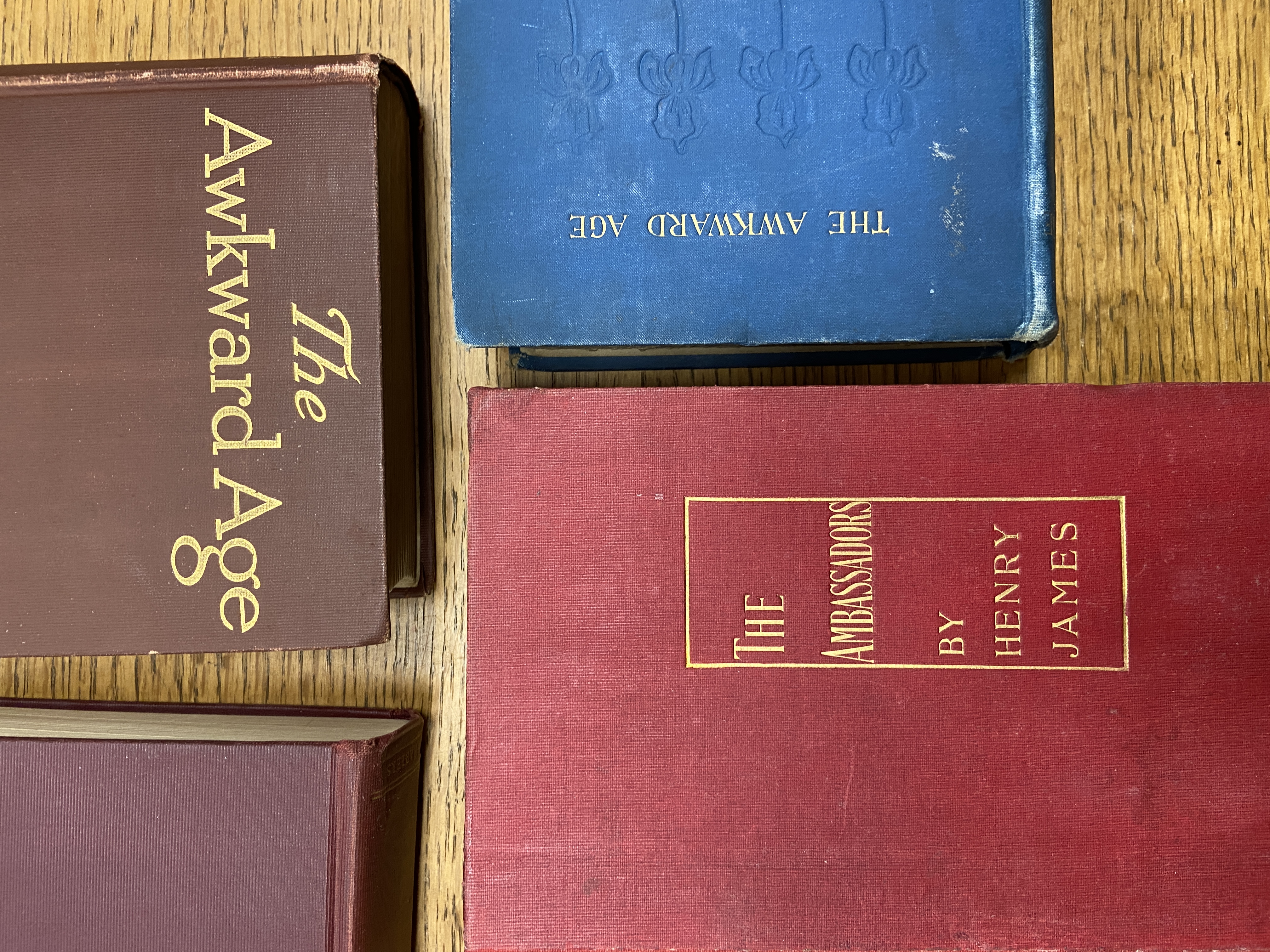 Close up detail of four books on a wooden table. Two copies of The Awkward Age (one bound in brown and one bound in blue) and The Ambassadors (one bound in dark red and one bound in light red).
