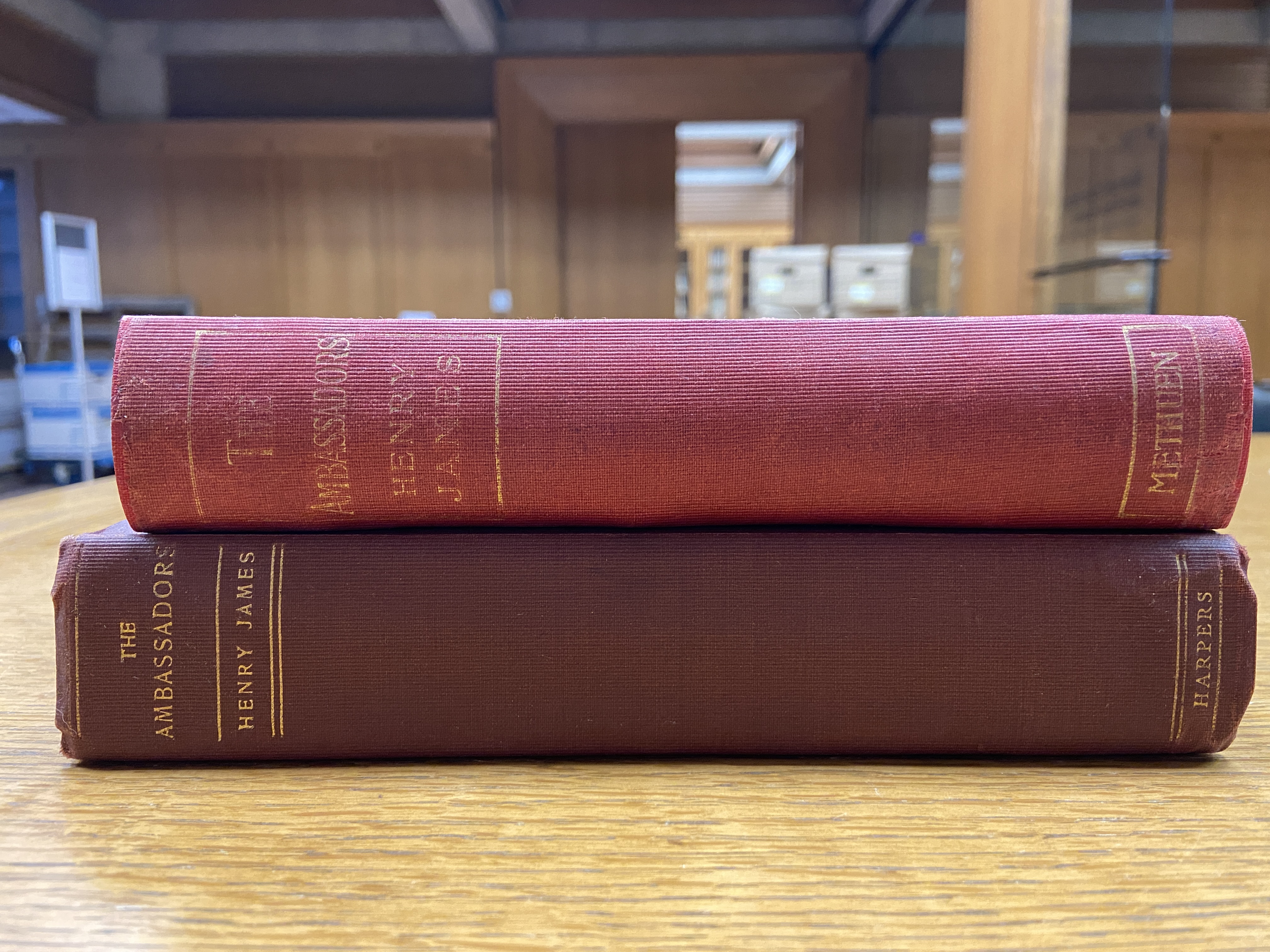 Two copies of The Ambassadors stacked on top of each other on a wooden table.The one on the bottom is bound in dark red and the one on the top is bound in light red. Both have gold text and embellishment.